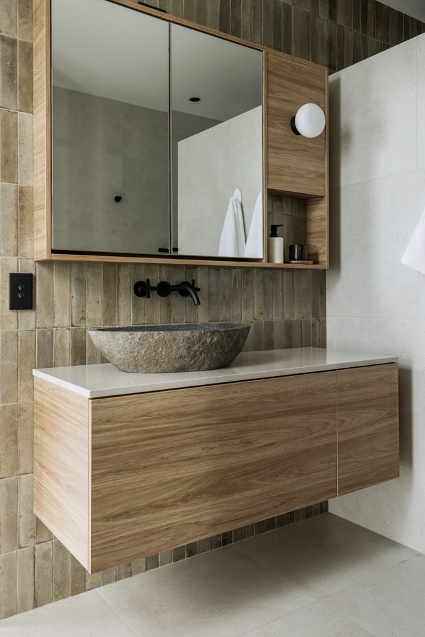 Contemporary bathroom vanity with a floating timber-grain cabinet, pale stone benchtop, textured vertical beige-grey wall tiles, and a rough natural stone vessel basin. A black wall-mounted tap and black-framed mirrored wall cabinet with integrated timber shelving complete the refined, earthy design.