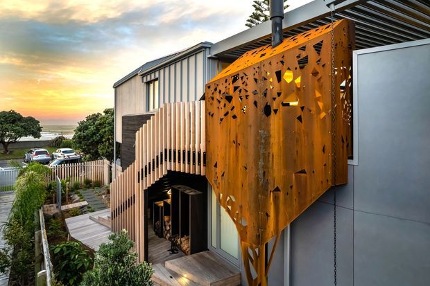 Exterior view of a contemporary multi-level residence on a sloped site, featuring a sculptural rust-orange perforated metal screen, vertical timber-look battens, dark cladding, a flat pergola roof, and an outdoor stair and garden path. Water and a suburban streetscape are visible in the distance.