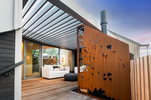 Contemporary residential outdoor living area with a timber deck, glass-lined living room, slatted pergola, and a large rusted corten-steel privacy screen featuring geometric cutouts. A white outdoor sofa and ottoman sit beneath the covered terrace, with dark horizontal exterior cladding and warm interior lighting visible beyond.