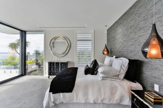 Contemporary bedroom with a large bed dressed in white bedding and black accents, set against a textured grey feature wall. Sculptural pendant lights hang beside the bed, while floor-to-ceiling glazing and a balcony or terrace provide bright outdoor views. The room also includes a dark bedside cabinet, wall-mounted artwork, and a narrow horizontal window with blinds.