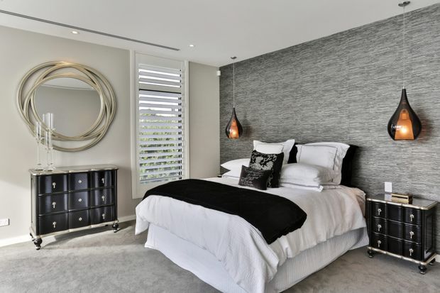 Contemporary bedroom with a large upholstered bed dressed in white bedding and a black throw, set against a textured grey feature wall. Matching dark bedside chests flank the bed, while a black dresser, sculptural circular mirror, pendant lights, plantation-style window shutters, and pale carpet complete the refined interior.