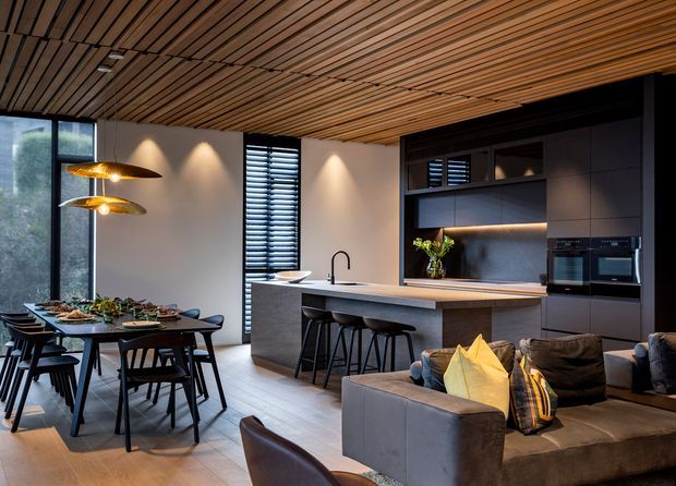 Open-plan contemporary kitchen, dining, and living area with a long island bench, dark built-in cabinetry, integrated appliances, black-framed glazing, and a dining table beside the kitchen. A slatted timber ceiling adds warmth and texture, while pendant lights illuminate the dining zone. The space has a refined, warm modern character with extensive built-in joinery and a neutral palette.