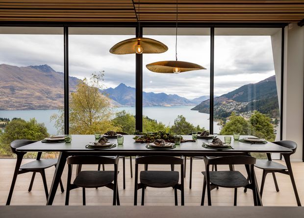 A contemporary dining room with a long dark dining table set for a meal and surrounded by modern chairs. Full-height black-framed glazing opens to expansive mountain and lake views, while a timber slatted ceiling and two sculptural pendant lights add warmth and visual interest.