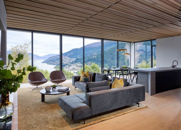 Open-plan contemporary living area with a grey sectional sofa, sculptural lounge chairs, round coffee table, dining table, and a minimalist kitchen island. Floor-to-ceiling glazing provides expansive mountain and water views, while a slatted timber ceiling and warm timber flooring add natural texture.