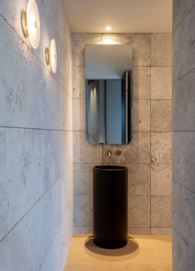 A compact, refined bathroom featuring pale textured stone-look wall tiles, a warm timber floor, a tall matte-black cylindrical pedestal basin with wall-mounted brass fixtures, and a rounded-rectangle mirrored cabinet. Soft integrated wall sconces and recessed ceiling lighting create a warm, moody atmosphere.