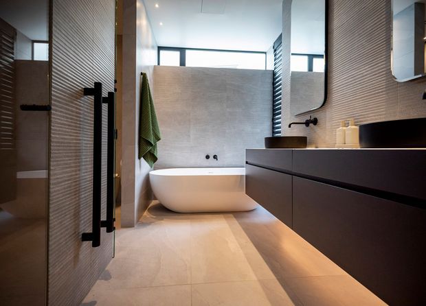 Contemporary bathroom with a freestanding white bathtub, floating dark vanity, integrated basin, wall-mounted black fixtures, large rounded-rectangle mirror, and textured horizontal wall tiles. Warm beige floor and wall finishes are balanced by black joinery and hardware, with natural light entering through high windows. A shower enclosure is visible at left.