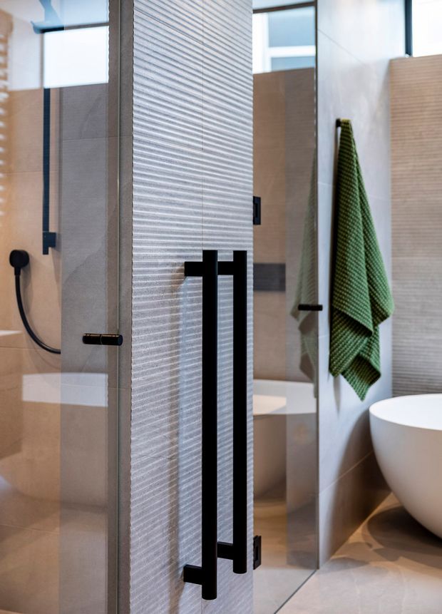 Close-up view of a contemporary bathroom featuring a frameless glass shower enclosure with a textured privacy panel and bold matte-black vertical hardware. A wall-mounted handheld shower, freestanding white bathtub, green textured towel, large mirror, and warm beige and grey tiled surfaces are visible.