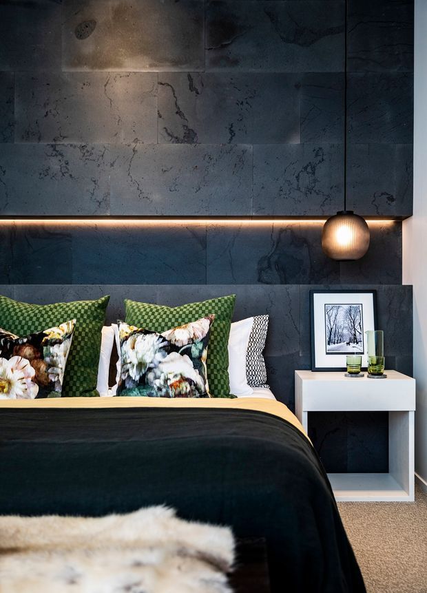 A moody, contemporary bedroom featuring a dark stone-effect tiled feature wall and integrated warm LED strip lighting behind the headboard. The bed is dressed in black and green textiles with patterned floral and geometric cushions, beside a minimalist white bedside table with framed artwork and glassware. A spherical pendant light adds a warm focal accent.