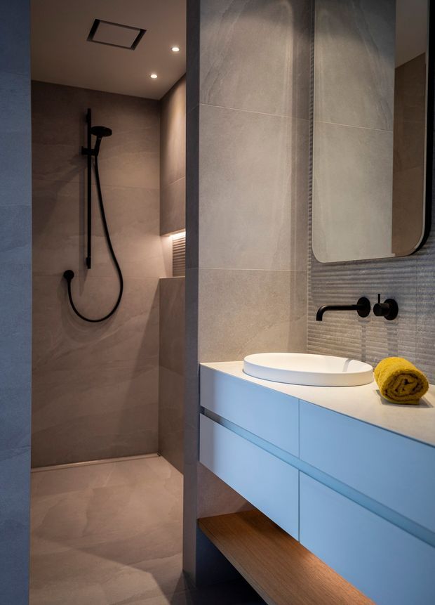 A compact contemporary bathroom featuring a walk-in shower with large warm-grey stone-look tiles, matte black shower and tapware, a floating pale blue vanity with a white vessel basin, and a mirrored wall cabinet. Integrated warm lighting and a timber open shelf add contrast and warmth.