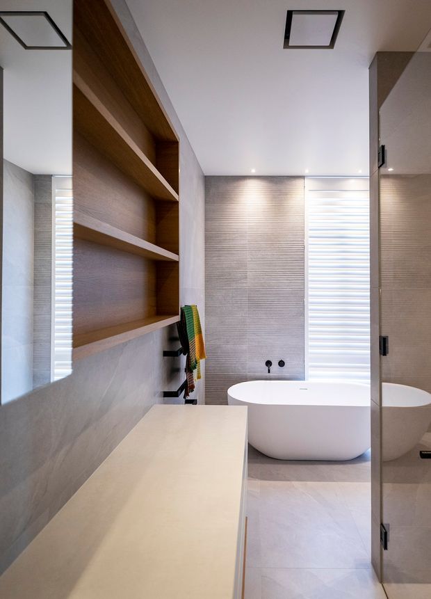 Contemporary bathroom featuring a freestanding white oval bathtub, pale large-format tiled surfaces, a textured horizontal feature wall, integrated open timber shelving, a light stone vanity, mirrored cabinetry, and bright recessed ceiling lighting. A partially visible shower or wet-area enclosure appears at the right.