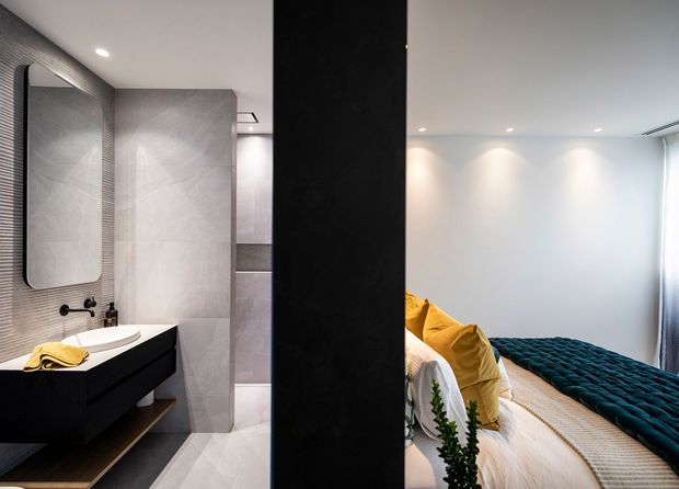 A contemporary interior view combining a minimalist bathroom and an adjacent bedroom. The bathroom features a floating dark vanity with a white basin, large light-grey wall and floor tiles, a frameless mirror, and recessed lighting. Through the opening, the bedroom includes a bed with white bedding, mustard-yellow cushions, and a dark teal textured throw, with clean white walls and soft natural light.