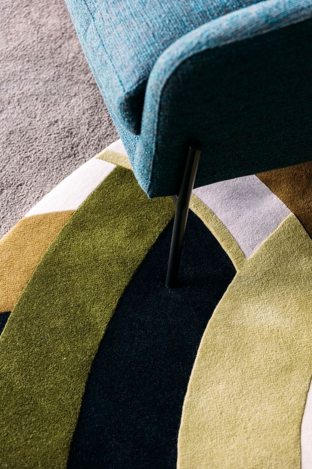 Close-up interior detail of a blue upholstered lounge chair with a slim dark metal leg, positioned on a bold abstract patterned rug. The rug features curved bands and geometric blocks in olive green, mustard, cream, pale lavender, and deep navy, with a neutral textured carpet visible at the edge.