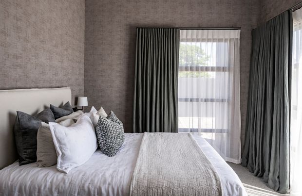 A refined bedroom with a large upholstered bed dressed in layered white, grey and patterned cushions. Textured taupe-grey walls, full-height dark charcoal curtains, sheer window coverings and soft natural light create a calm, monochromatic interior.