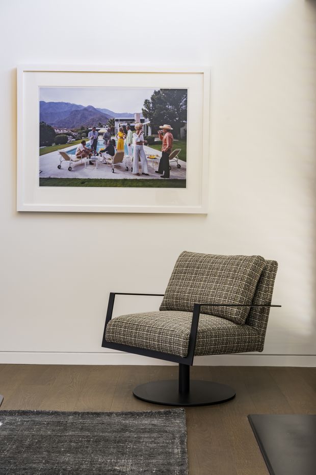 A refined interior vignette featuring a contemporary upholstered lounge chair with a black metal frame and pedestal base, positioned against a warm off-white wall. A framed photographic artwork hangs above the chair, while dark timber flooring and a charcoal textured rug add contrast. The space is minimalist and carefully styled, with soft neutral tones and clean lines.