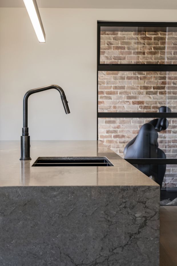 Close-up view of a contemporary kitchen island with a dark stone waterfall edge, integrated rectangular sink and matte black gooseneck tap. A black-framed internal window or partition reveals an exposed brick wall and black sculptural pendant lighting beyond. The palette is neutral and industrial-leaning, with warm grey stone, black metal and brick accents.