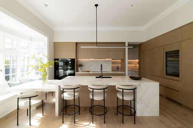 Bright, refined contemporary kitchen with a large pale stone island, three black-framed upholstered bar stools, integrated timber cabinetry, a stone backsplash, and a built-in dark appliance wall. A dining nook with curved seating sits beside tall traditional-style windows, while a linear pendant light and concealed warm cabinetry lighting add contrast.