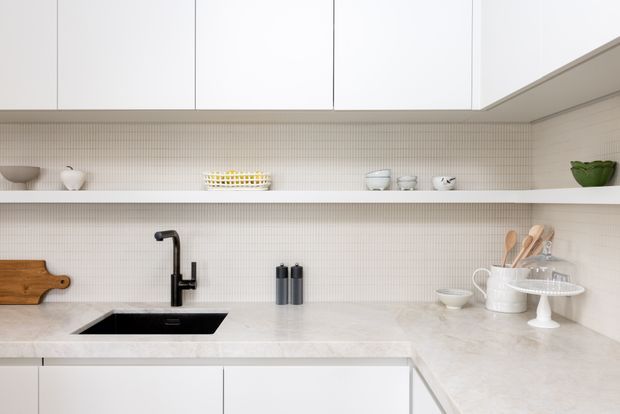 Bright, minimalist contemporary kitchen with white flat-panel cabinetry, open shelving, a pale stone benchtop, fine-grid tiled splashback, and a black undermount sink with matching tap. Neutral ceramics, timber utensils, and a small green bowl provide restrained accents.