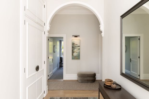 A bright, refined residential entry hall viewed from inside, featuring a large arched opening with decorative plaster detailing, white paneled cabinetry or doors, light neutral walls, timber flooring, layered rugs, a dark timber console, and a large framed mirror. A secondary hallway and doorway are visible beyond the arch.