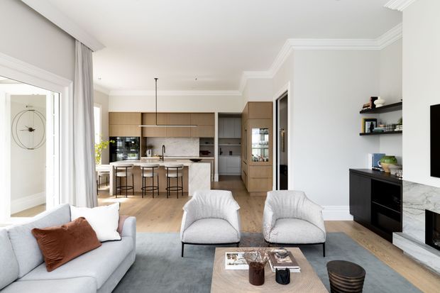 Bright, refined open-plan living area with a pale upholstered sofa and two armchairs arranged around a low coffee table, opening directly into a contemporary kitchen with a central island and stools. The room has light timber flooring, pale grey walls, white decorative ceiling cornices, full-height curtains, built-in timber cabinetry, dark shelving and cabinetry, and a marble-look fireplace surround. A glazed door and tall windows bring in abundant natural light.