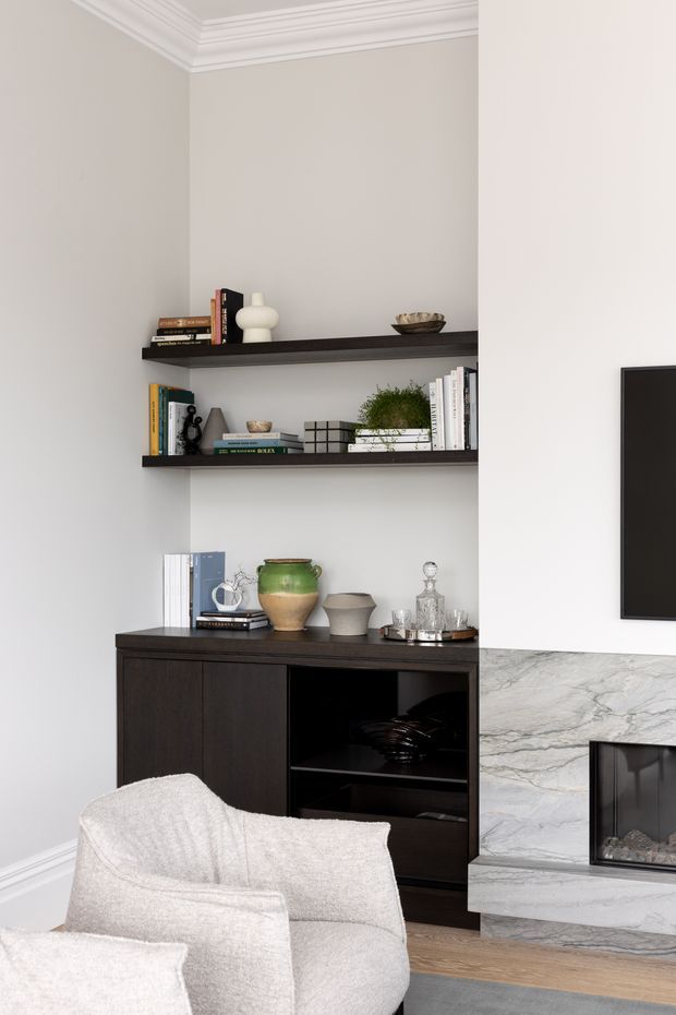 A refined living room vignette featuring dark built-in shelving and cabinetry set into a light neutral wall, styled with books, ceramics, greenery, and glassware. A marble-clad fireplace and wall-mounted television are visible to the right, with pale upholstered armchairs in the foreground. White decorative ceiling cornices and light timber flooring complete the room.