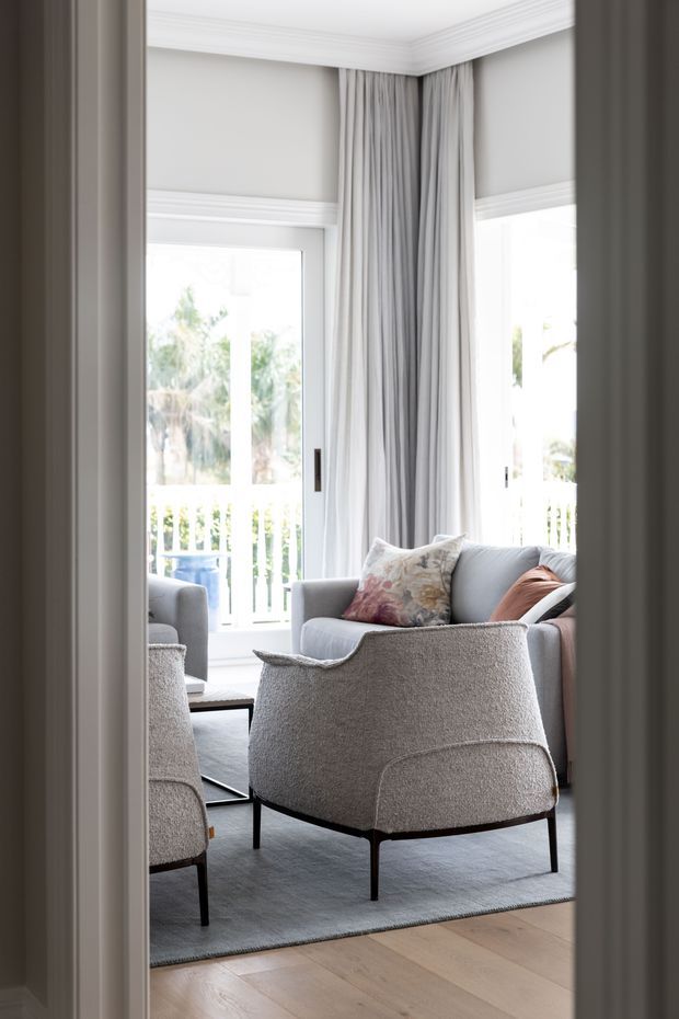 A bright, refined living room viewed through a doorway, featuring pale grey upholstered lounge chairs and sofa, a large soft grey rug, light timber flooring, tall windows with sheer and full-length grey curtains, and traditional white crown moulding. The room opens onto a balcony or veranda with leafy views outside.