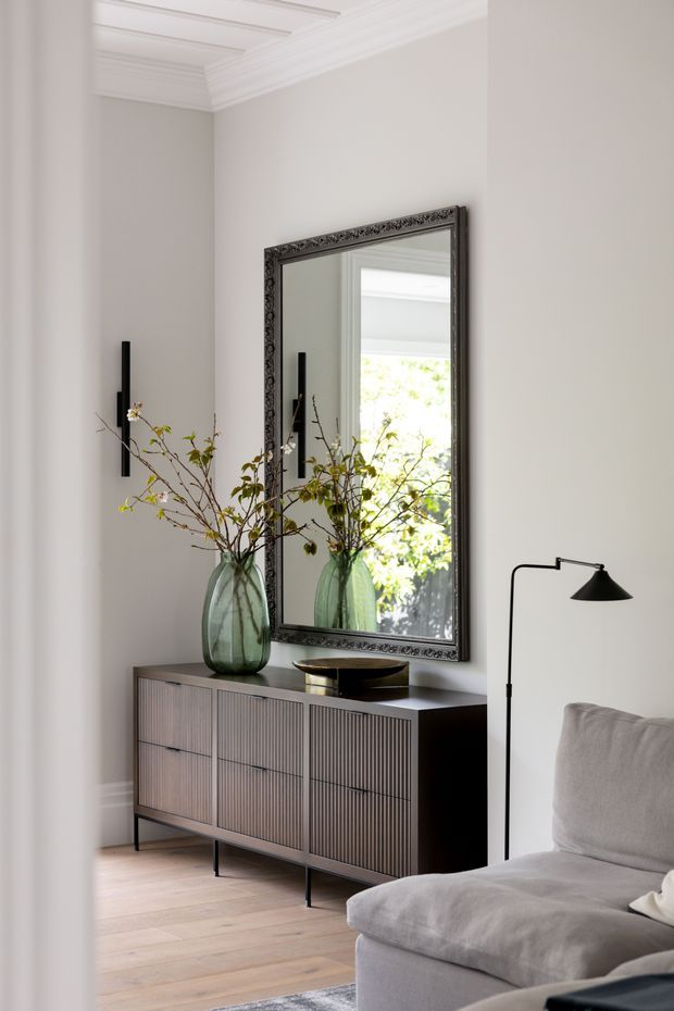 A refined living room corner featuring a dark fluted sideboard, large ornate black-framed mirror, sculptural glass vase with branches, black wall sconces, and a grey upholstered sofa. The room has pale neutral walls, light timber flooring, and detailed white ceiling cornices, creating a calm contemporary-classic atmosphere.