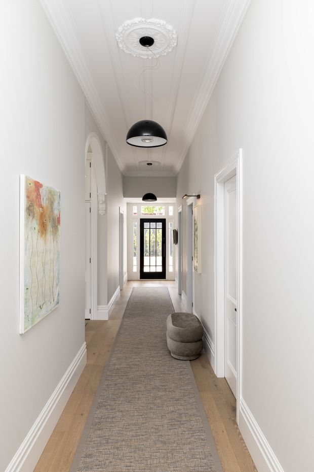 A bright, narrow residential hallway with white paneled walls, ornate ceiling moulding and medallion, black pendant lights, pale timber flooring, and a long neutral runner rug. White-trimmed doorways line both sides, with a black-framed glazed entry door at the far end. A small rounded upholstered stool and abstract artwork add understated contemporary detail.