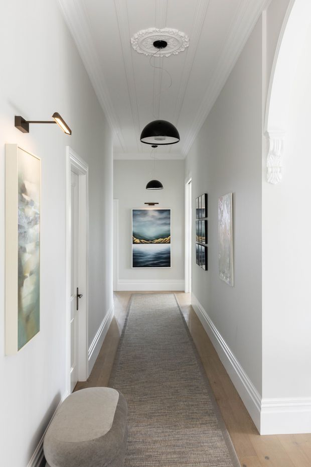Bright, narrow residential hallway with pale grey walls, white decorative trims, a detailed pressed-panel ceiling and ornate ceiling medallion. Black pendant lights run along the corridor, with contemporary artwork on both walls, a long neutral runner, timber flooring, and a rounded upholstered bench near the foreground. A large arched opening and classical mouldings add architectural character.