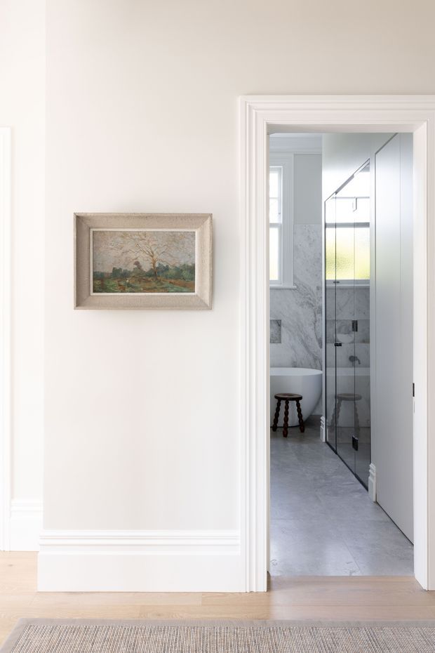 A bright, refined interior view from a hallway into a marble-finished bathroom. The foreground has pale timber flooring, white painted walls with decorative skirting and trim, a framed landscape artwork, and a neutral woven rug. Through the doorway, the bathroom includes a freestanding white bathtub, large grey-veined marble wall and floor surfaces, and a black-framed glass shower enclosure.