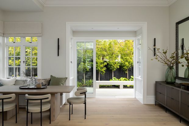 Refined contemporary dining and living area with pale timber flooring, a light wood dining table, upholstered dining chairs, and a dark ribbed sideboard. The room features white-painted walls, ornate layered ceiling cornices, tall divided-light windows, and a large glazed opening with white French doors leading to a small landscaped courtyard. A large framed mirror, sculptural wall lighting, and a vase of branches complete the calm, light-filled interior.