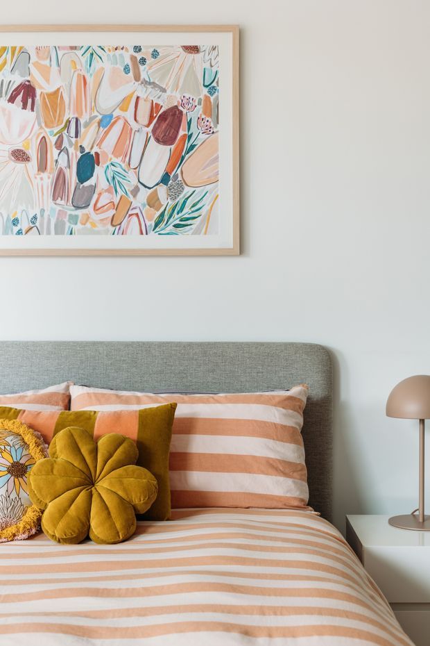 A bright, styled bedroom vignette featuring a pale grey upholstered bedhead, striped peach and cream bedding, layered decorative cushions including a mustard velvet flower cushion, and a minimalist bedside table with a rounded brass-toned lamp. A colourful botanical-style framed artwork hangs above the bed against a soft off-white wall.