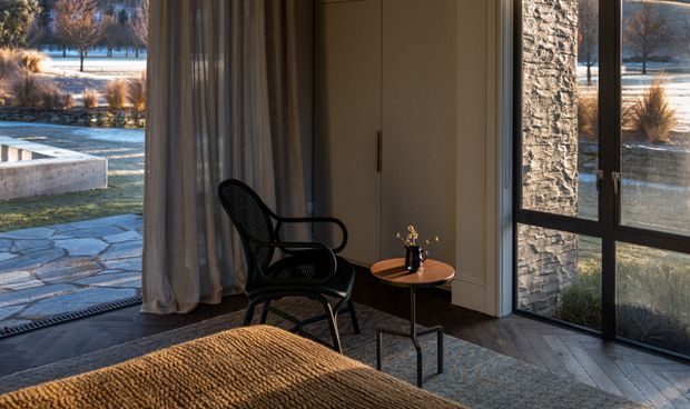 A calm, refined interior corner with a black woven lounge chair beside a small round timber side table, heavy floor-length curtains, a built-in beige storage cabinet, and dark-framed floor-to-ceiling glazing. A textured upholstered or woven foreground surface and grey-toned flooring complete the room. Through the glazing, a stone-paved terrace and landscaped, wintery exterior are visible.