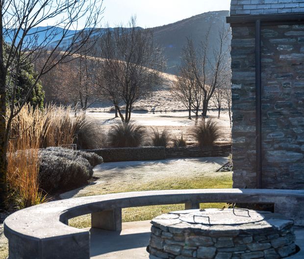 Exterior view of a landscaped mountain property in winter, featuring a curved concrete seating wall around a circular stacked-stone fire pit or water feature, ornamental grasses, clipped shrubs, leafless trees, and a stone-clad building edge. Snow-dusted hills and frosty lawn form the background.