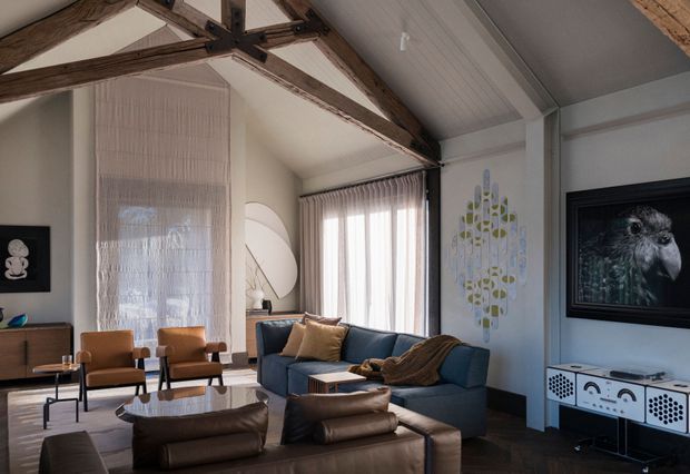 A refined living room with a high pitched ceiling and exposed rustic timber trusses. The space features a deep blue sofa, tan leather lounge chairs and ottomans, dark timber flooring, sheer curtains, a sculptural pendant, framed artwork, and a patterned sideboard. The palette is warm and earthy with a relaxed, sophisticated character.