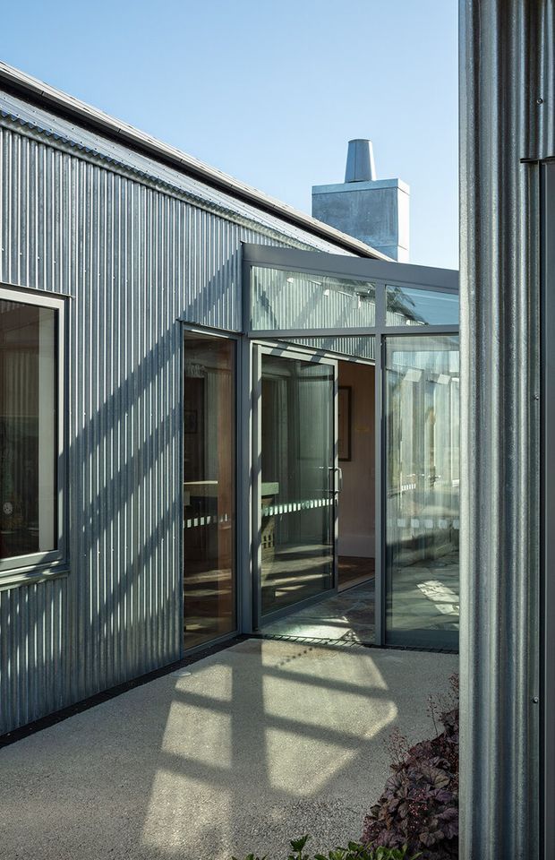 Exterior view of a contemporary corrugated-metal-clad building with a glazed aluminium-framed entry or connecting vestibule. The image shows a pitched roof, metal chimney or rooftop vent, full-height glass doors and windows, and a concrete or gravel-like paved approach with strong angular sunlight and shadows.