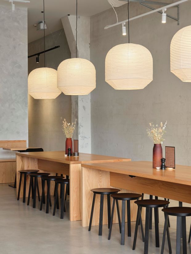 A contemporary café or dining venue with long pale timber communal counters and black stools. Large ribbed spherical pendant lights hang over the counters, while exposed concrete-look walls, visible track lighting, and simple black metal details create a refined industrial atmosphere. Small vases and menu stands provide restrained decorative accents.