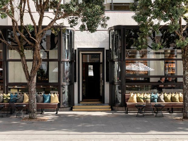 Street-facing facade of a refined multi-storey commercial or hospitality building, framed by mature trees. The light textured exterior is articulated with dark metal-framed glazing, tall curved projecting window bays, and a central recessed entry. Built-in seating with colourful cushions lines the frontage, creating an inviting outdoor threshold along the paved sidewalk.