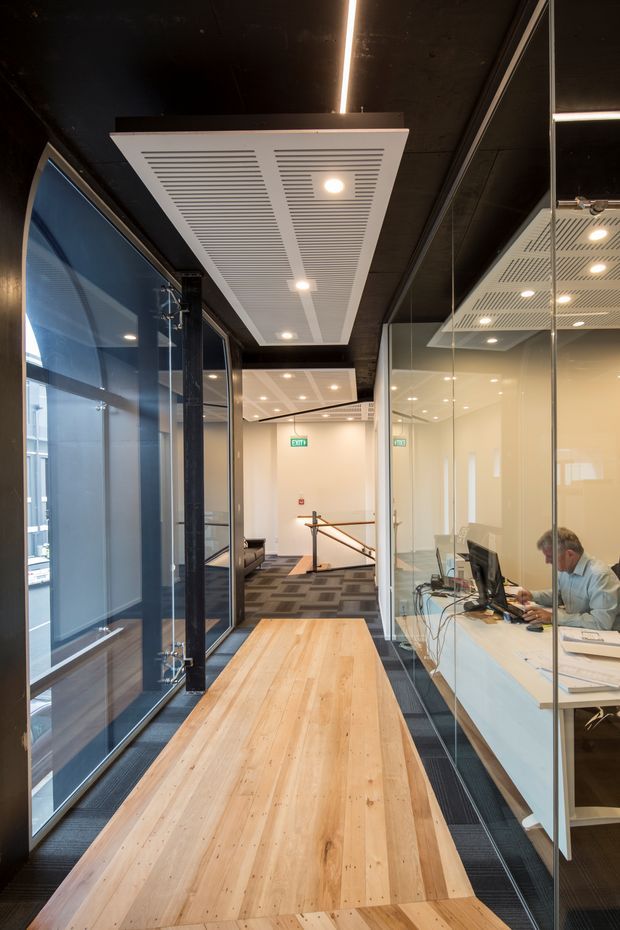 A narrow contemporary commercial office corridor with full-height glazed meeting or office partitions on both sides. The space features a long light timber floor and matching timber ledge, dark exposed ceiling and structural framing, linear LED lighting, recessed spotlights, patterned carpet in the adjoining area, and a visible workstation behind glass. A stair and office circulation area are visible toward the rear.