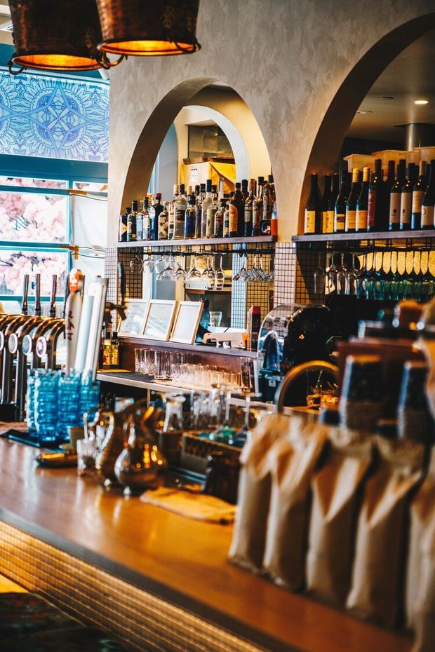 A richly styled commercial bar interior with arched plaster openings, dark metal shelving, extensive liquor and wine displays, hanging glassware, beer taps, and a polished timber-toned bar counter. Decorative blue patterned wall art and warm pendant lighting add colour and atmosphere.