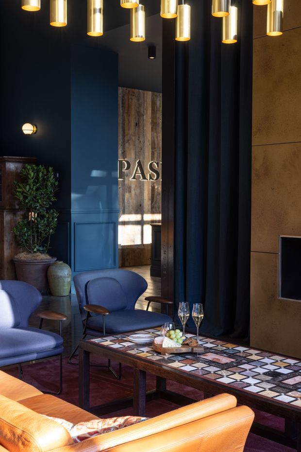 A moody, sophisticated lounge or hospitality-style interior with deep navy walls and curtains, warm brass and cylindrical pendant lights, dark timber and brown finishes, and a striking rectangular coffee table with a multicoloured patterned tiled surface. Blue upholstered lounge chairs, a caramel leather sofa, a potted plant, a drinks arrangement, and a partial fireplace or wall feature create a refined, intimate atmosphere. A timber-clad doorway or feature wall is visible in the background.