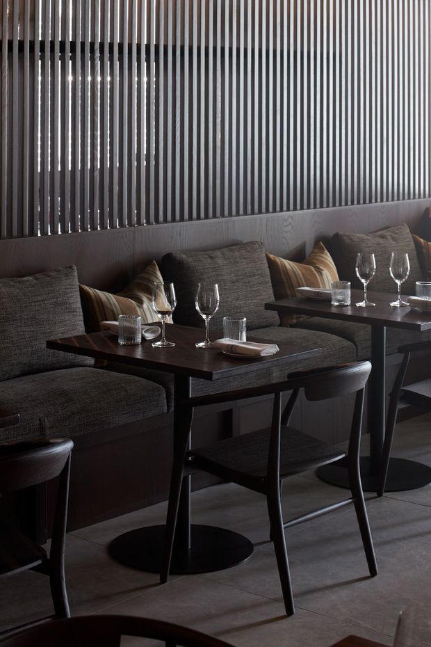A moody, refined restaurant dining area with dark upholstered banquette seating, small dark timber or stained tables, black pedestal bases, and matching dining chairs. Warm striped cushions and carefully set wine glasses, tumblers, plates, and cutlery create an intimate hospitality setting. A vertical dark slatted screen forms the backdrop above a timber-look wall panel, with large grey floor tiles below.