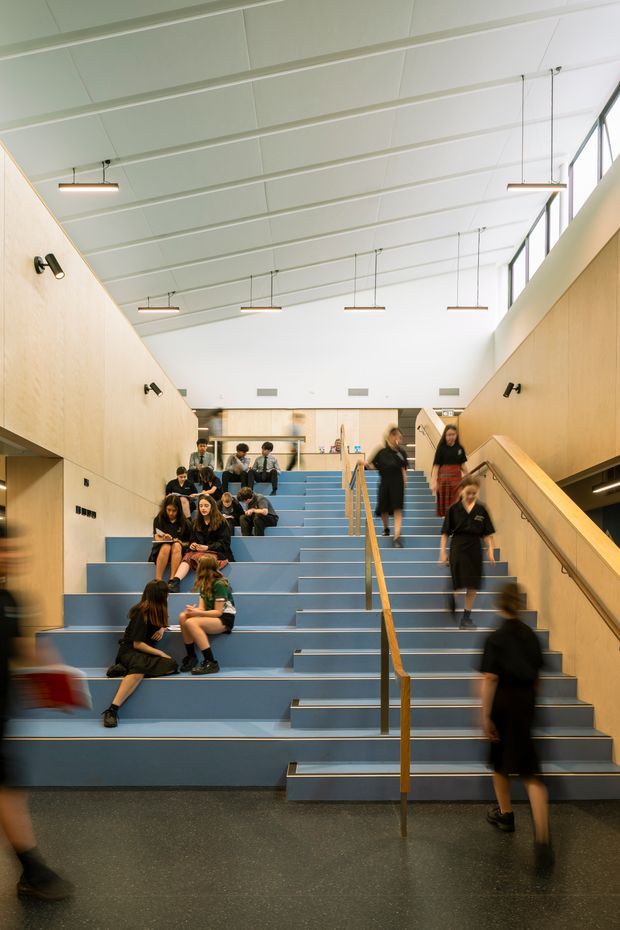 A bright contemporary institutional interior featuring a broad blue stepped stairway with integrated seating, wood-panelled side walls, timber handrails, and multiple suspended linear lights. Students and people occupy the steps and move through the space, which appears to function as a social circulation or gathering area. A high sloping ceiling with clerestory glazing brings in abundant natural light.