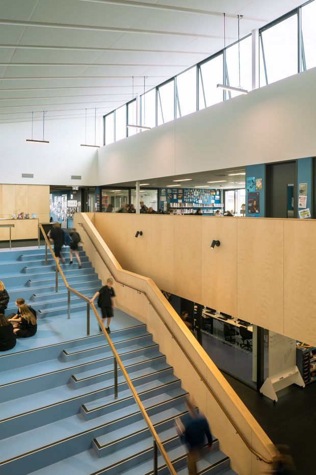A bright multi-level educational interior, likely a school or library, viewed from an upper landing over a broad blue-painted staircase. Pale timber-clad balustrades and partition walls surround a lower-level study or library area, with extensive internal glazing, high clerestory windows, suspended linear lights, and a white tiled ceiling. People are moving on the stairs and seated below.