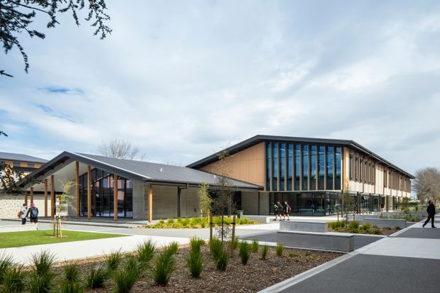Exterior architectural photograph of a contemporary institutional or commercial building arranged around landscaped paved forecourts. The complex features low-pitched metal roofs, warm timber-toned wall cladding, extensive dark-framed glazing, vertical sun-shading fins, and covered entrance canopies. Young trees, ornamental grasses, concrete seating or retaining elements, and pedestrian paths occupy the foreground.