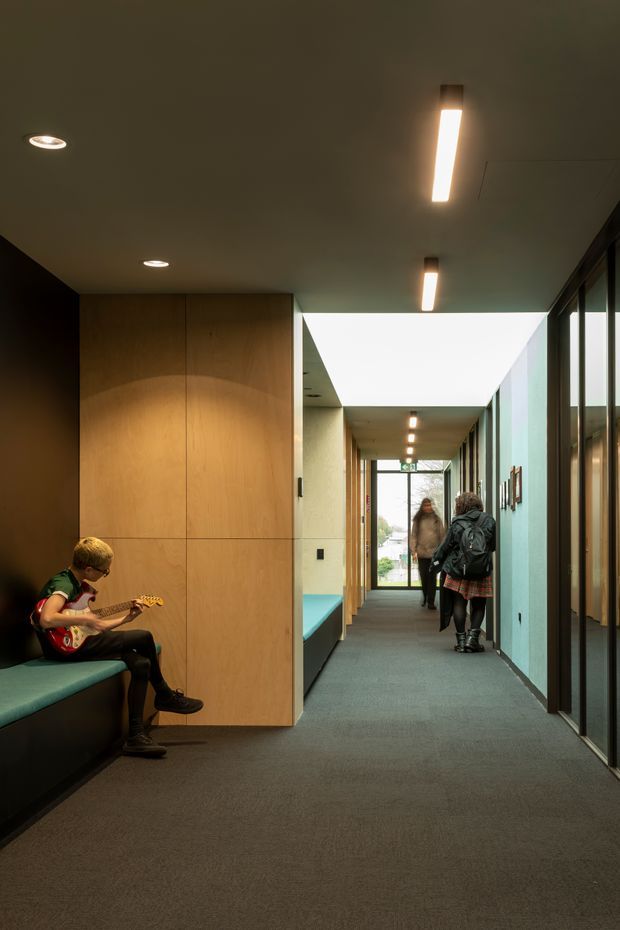 A long, contemporary interior corridor with dark carpet flooring, warm timber-lined built-in cabinetry and bench seating, black-framed glazed partitions on the right, and a pale ceiling with linear recessed lighting. A turquoise upholstered bench runs along the left wall, where a child is seated playing an electric guitar. The corridor leads toward a glazed exit with daylight beyond, and several people are visible farther down the hall.