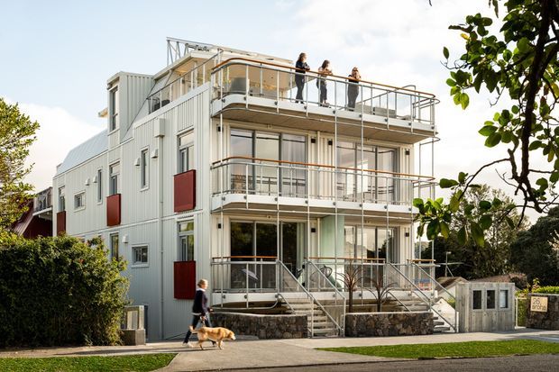 A multi-storey contemporary residential building viewed from the street, featuring pale corrugated metal cladding, extensive stacked balconies with slim metal balustrades, large glazed doors and windows, and external stair access. A landscaped hedge and front path frame the facade.