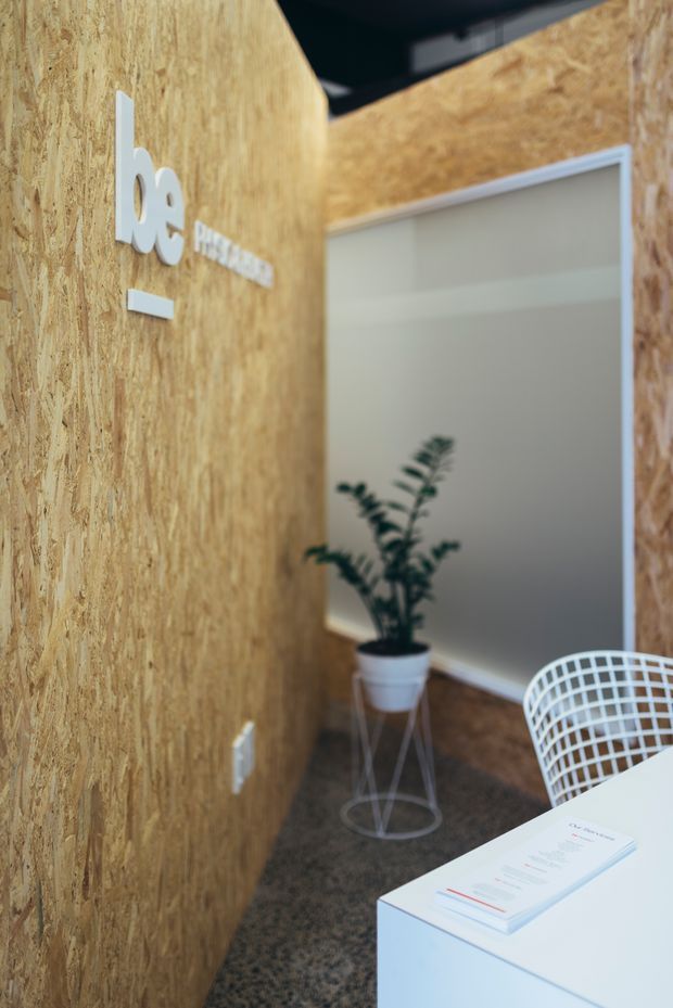 A compact modern commercial reception or waiting area featuring exposed OSB wood-panel walls, a white wall-mounted logo, a potted plant, white wire-frame seating, and a white reception desk with printed materials. The space has dark carpet flooring and a partially visible dark ceiling.