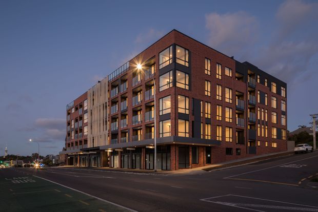 Dusk exterior view of a contemporary multi-storey apartment building on an urban street corner. The building features a prominent red-brown brick facade, large glazed windows, recessed balconies, dark metal framing, and contrasting light vertical facade panels. Ground-level commercial or lobby glazing fronts the street, with road markings and street lighting visible.