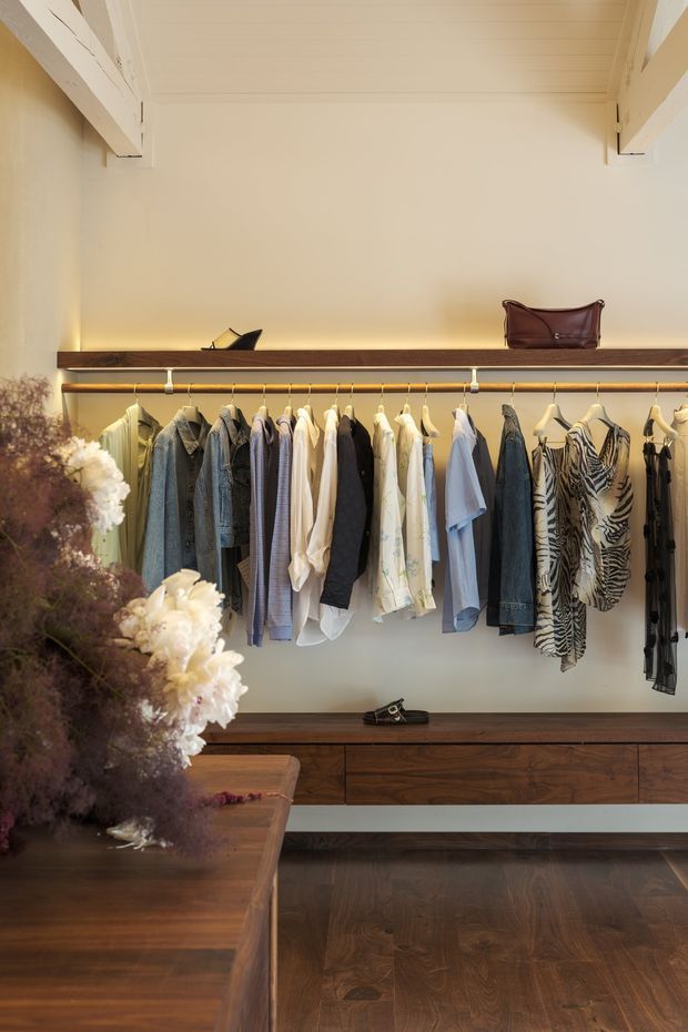 A warm, refined walk-in wardrobe with open hanging rails, dark timber drawer cabinetry, and a matching timber island or console in the foreground. Cream walls and a paneled ceiling are accented by integrated warm lighting, while clothing and handbags are arranged along the built-in storage. A large floral arrangement adds a decorative foreground element.
