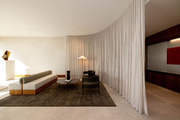 Refined contemporary living room with a low modular sofa, dark round coffee table, lounge chair, sculptural floor lamp and large textured area rug. A full-height curved sheer curtain forms a soft partition around the seating area, while a dark timber built-in joinery wall and red artwork or panel are visible beyond.